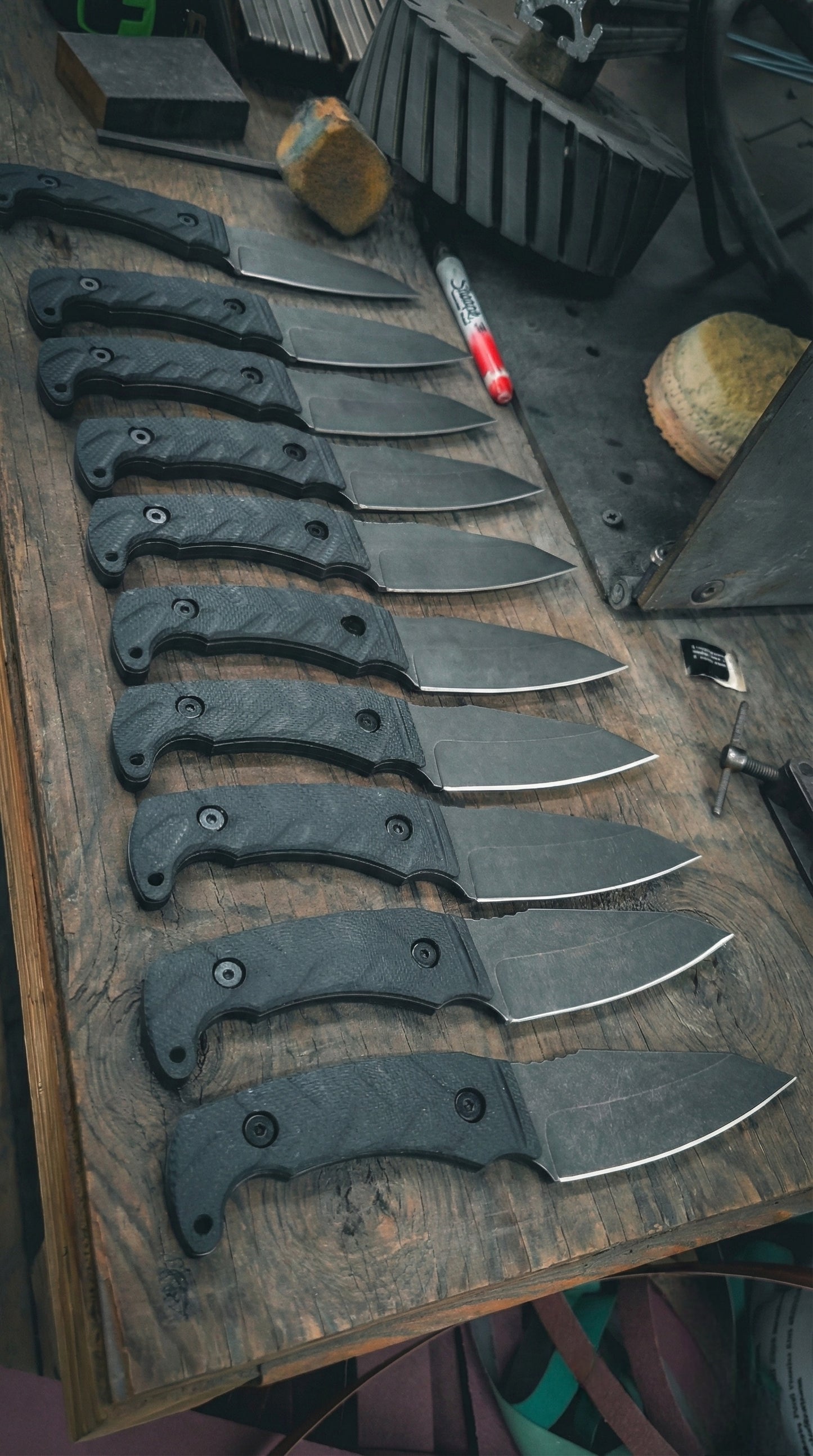 Row of black-handled knives on a wooden surface with tools in the background